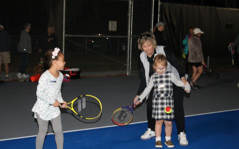 Genesis Wormely (from left) looks on as Abigayle Chadwick gets help in hitting a tennis ball from Dolly Robinson, Lake City Columbia Community Tennis Association president, during the ceremony opening The Tennis Courts at Youngs Park on Monday afternoon. (TONY BRITT/Lake City Reporter)