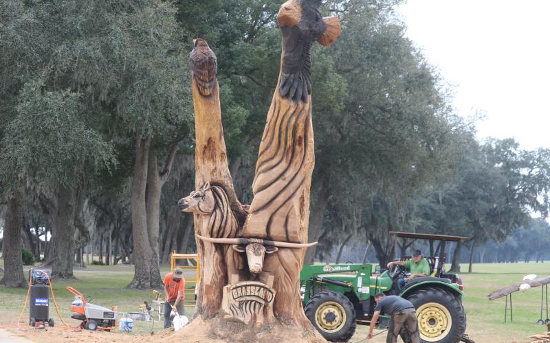 Kevin Treat (from left) and Josh Miller clean up around the custom wood carving they created with chainsaws for Carl Allison, who is seated on the tractor. (TONY BRITT/Lake City Reporter)