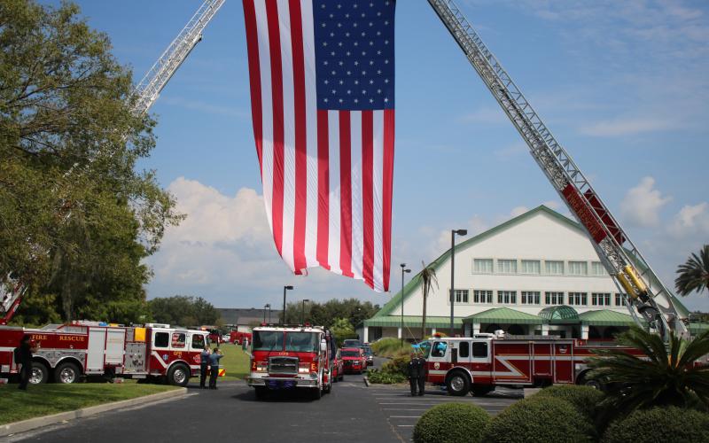 The procession, with Fire Chief Randy Burnham carried on the back of a Lake City fire truck, leaves Christian Fellowship Church on Friday, driving below a giant American flag. (TONY BRITT/Lake City Reporter)