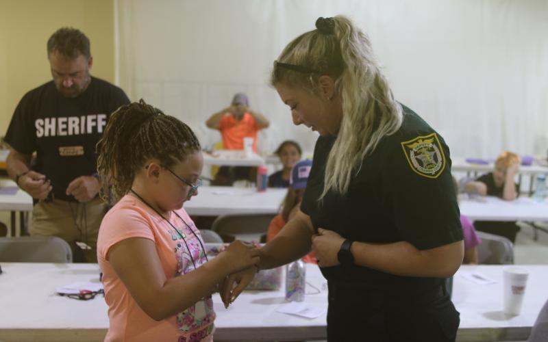 Giana Rivera (left) ties a friendship bracelet around Columbia County Sheriff’s Office Deputy Stormi McAdams’s wrist during the Harmony In The Streets camp. (TONY BRITT/Lake City Reporter)