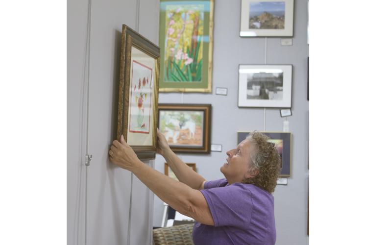 Janet Hans, an artist and a volunteer jewelry-making instructor at the Gateway Art Gallery, hangs artwork for the Gallery’s Christmas in July reception today. (TONY BRITT/Lake City Reporter)