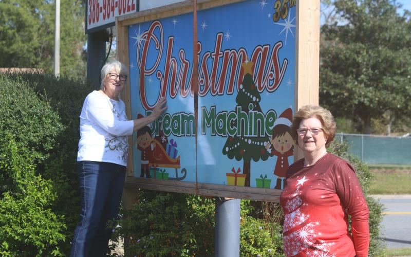 Meally Jenkins (from left), Christmas Dream Machine founder and director and Janet Stanford, Christmas Dream Machine treasurer and secretary, pose near organization’s sign in Country Club Plaza on Tuesday morning. (TONY BRITT/Lake City Reporter)
