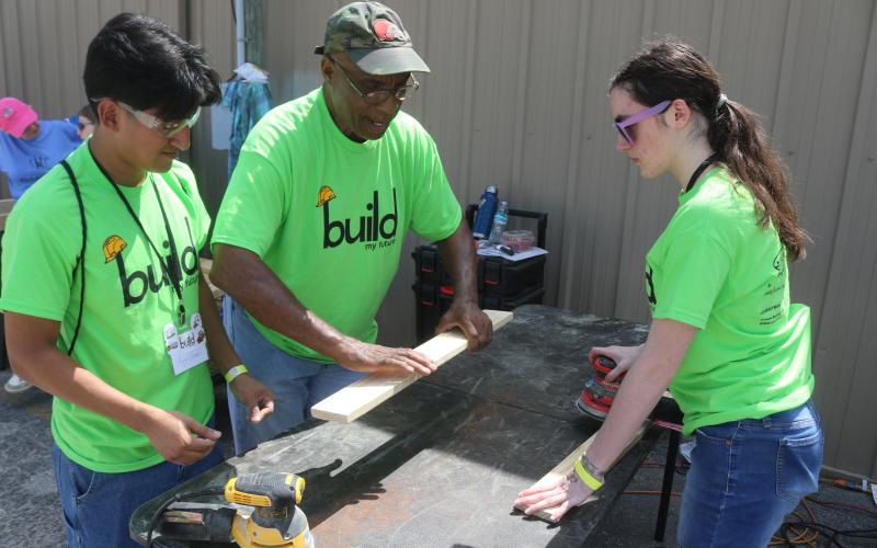Angel Gomez, a Columbia High School student, gets tips on sanding wood from Monty Ashby, as Gomez and Kiley Morrison sand wood to make beds for the Sleep in Heavenly Peace program. The students manufactured the beds during the Build My Future event on Wednesday morning at the Florida Gateway Fairgrounds. (TONY BRITT/Lake City Reporter)