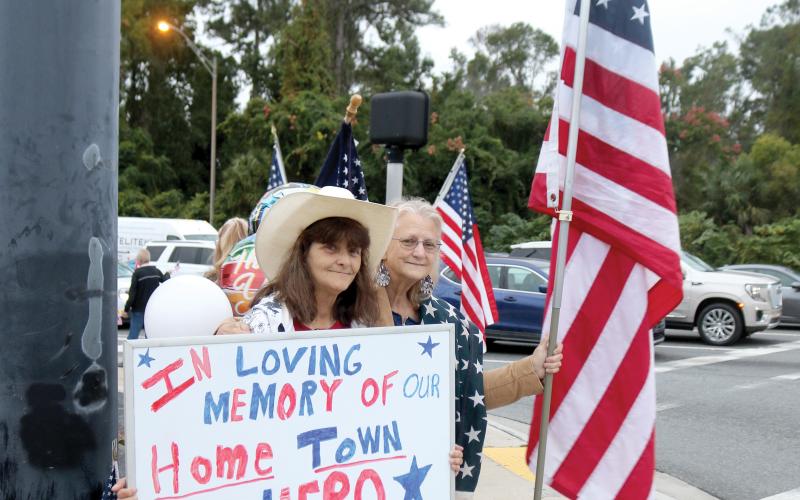 Bonnie Smith (from left) and Doreen Waters, the late Terry Hancock’s sisters, hold flags and a sign for motorists in honor of their brother on Wednesday morning. (TONY BRITT/Lake City Reporter)