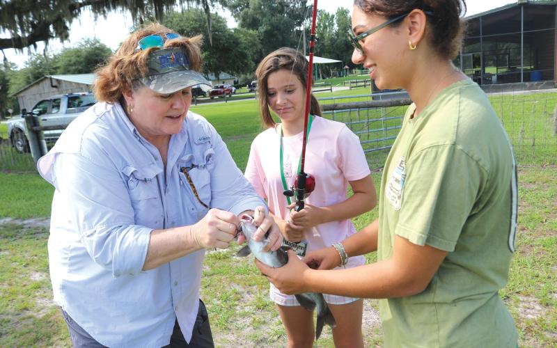 Paulette Tomlinson (from left), a Columbia County Extension Service employee, takes the hook out of a catfish for Madelyn Bullock, 13, with help from Paola Ortiz, a 4-H summer camp volunteer. (TONY BRITT/Lake City Reporter)