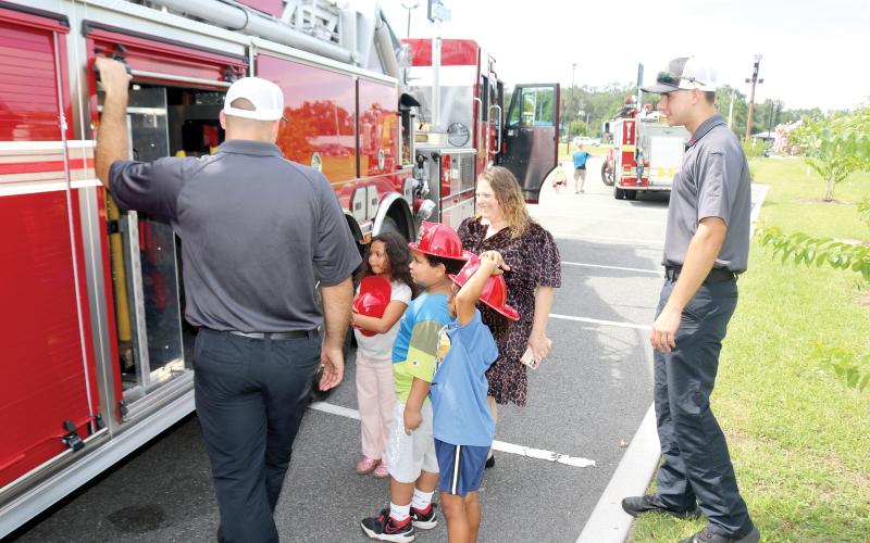 Lake City Fire Department firefighter Chase Hancock (from left) lets Ryker Paris, 7, Jeremiah Williamson, 7, Ariella Paris, 7 and Melissa Angeles look at the equipment firefighters use on emergency calls during the Firefighters and Cones event. (TONY BRITT/Lake City Reporter)