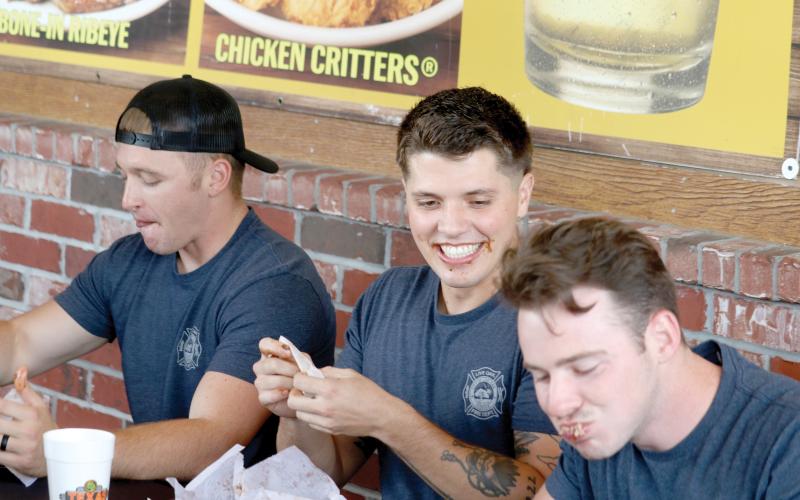 Live Oak firefighter Zach Yarnell (center) grins as Cody Byers (right) has a mouthful of ribs during the contest Tuesday. Kessler Mallory (left) also participated for the Live Oak Fire Department team. (TONY BRITT/Lake City Reporter)