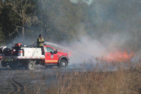 An unidentified firefighter with Columbia County Fire Rescue stands on the back of a brush truck while spraying water on a brush fire Tuesday afternoon. (TONY BRITT/Lake City Reporter)