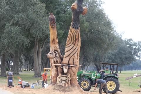 Kevin Treat (from left) and Josh Miller clean up around the custom wood carving they created with chainsaws for Carl Allison, who is seated on the tractor. (TONY BRITT/Lake City Reporter)