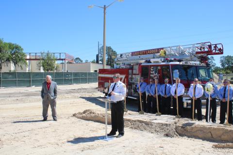 Stephen Witt (far left) looks on as Josh Wehinger, Lake City Fire Department Chief, addresses the audience during a groundbreaking ceremony for Lake City Fire Station 2 with a group of firefighters in the background.