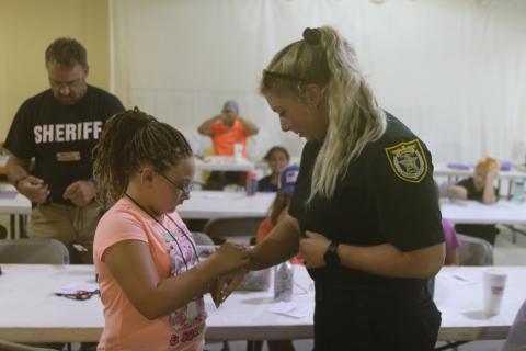 Giana Rivera (left) ties a friendship bracelet around Columbia County Sheriff’s Office Deputy Stormi McAdams’s wrist during the Harmony In The Streets camp. (TONY BRITT/Lake City Reporter)