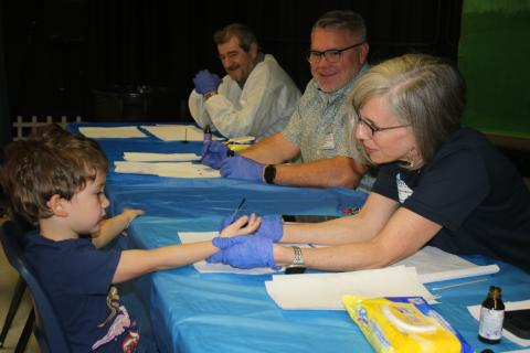 Grason Montero, 4, allows Rotary Club of Lake City member Margot Maranto to paint his pinkie purple Friday morning at Westside Elementary School as fellow Rotarians Ron Turbeville and Fred Koberlein look on with smiles. (TONY BRITT/Lake City Reporter)