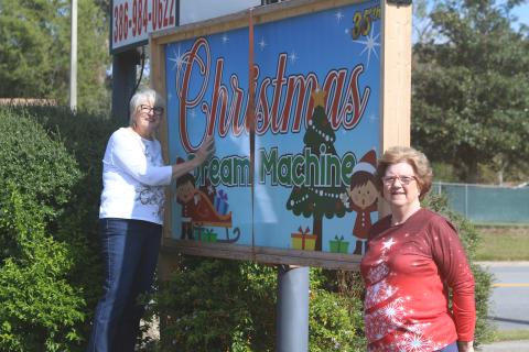 Meally Jenkins (from left), Christmas Dream Machine founder and director and Janet Stanford, Christmas Dream Machine treasurer and secretary, pose near organization’s sign in Country Club Plaza on Tuesday morning. (TONY BRITT/Lake City Reporter)