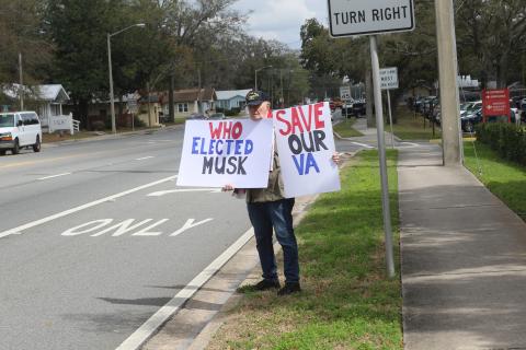 Charles Trowbridge, a U.S. Navy veteran and Fort White resident, holds up signs outside the Lake City VA Medical Center on Monday protesting the Department of Government Efficiency’s planned cutting of 80,000 Veterans Affairs employees. (TONY BRITT/Lake City Reporter)
