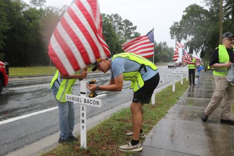 Chase Piesel (right) uses a drill to secure a flag to a cross on Saturday morning as volunteers erected flags in honor of veterans and first responders. (TONY BRITT/Lake City Reporter)