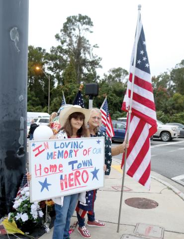 Bonnie Smith (from left) and Doreen Waters, the late Terry Hancock’s sisters, hold flags and a sign for motorists in honor of their brother on Wednesday morning. (TONY BRITT/Lake City Reporter)