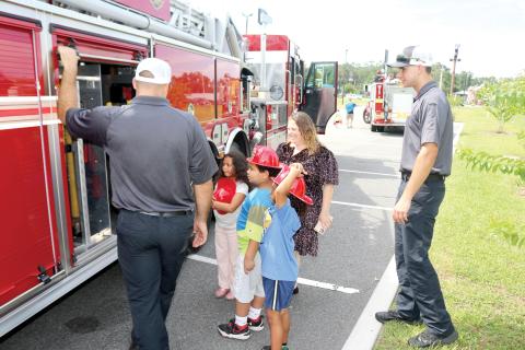 Lake City Fire Department firefighter Chase Hancock (from left) lets Ryker Paris, 7, Jeremiah Williamson, 7, Ariella Paris, 7 and Melissa Angeles look at the equipment firefighters use on emergency calls during the Firefighters and Cones event. (TONY BRITT/Lake City Reporter)