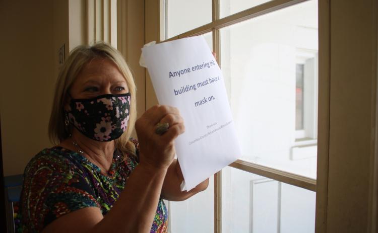 Marsha Wallace, Columbia County School Board office information specialist, places a sign on the central office’s door notifying the general public they must wear masks before entering the building. (TONY BRITT/Lake City Reporter)
