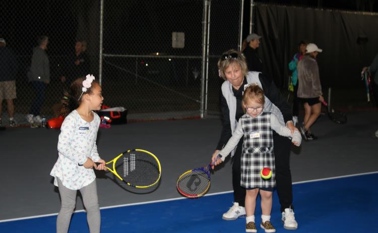 Genesis Wormely (from left) looks on as Abigayle Chadwick gets help in hitting a tennis ball from Dolly Robinson, Lake City Columbia Community Tennis Association president, during the ceremony opening The Tennis Courts at Youngs Park on Monday afternoon. (TONY BRITT/Lake City Reporter)