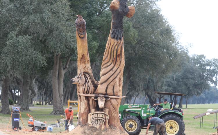 Kevin Treat (from left) and Josh Miller clean up around the custom wood carving they created with chainsaws for Carl Allison, who is seated on the tractor. (TONY BRITT/Lake City Reporter)