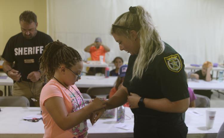 Giana Rivera (left) ties a friendship bracelet around Columbia County Sheriff’s Office Deputy Stormi McAdams’s wrist during the Harmony In The Streets camp. (TONY BRITT/Lake City Reporter)