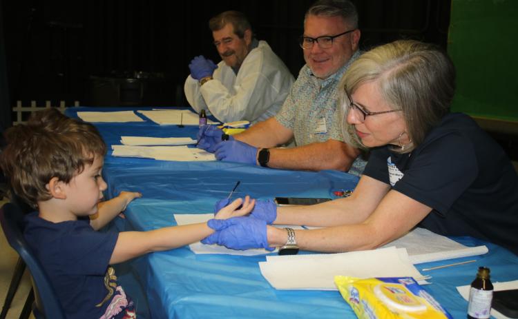 Grason Montero, 4, allows Rotary Club of Lake City member Margot Maranto to paint his pinkie purple Friday morning at Westside Elementary School as fellow Rotarians Ron Turbeville and Fred Koberlein look on with smiles. (TONY BRITT/Lake City Reporter)