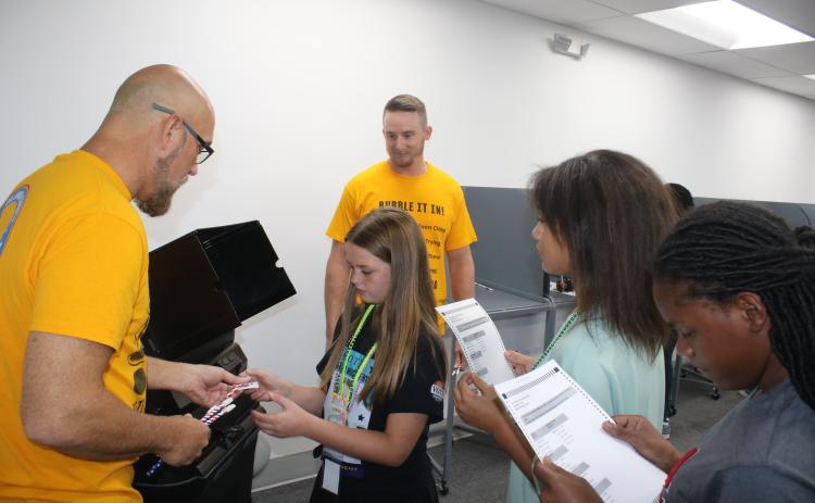 Columbia County Supervisor of Elections Office employee Dennis Chauncey hands a sticker to an Eastside Elementary School fifth grader after she voted in the school’s election Tuesday. Other students wait in line with their ballots. (TONY BRITT/Lake City Reporter)