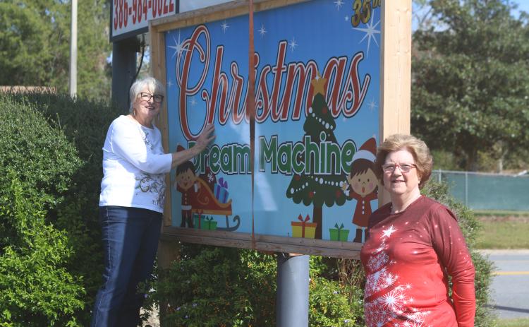 Meally Jenkins (from left), Christmas Dream Machine founder and director and Janet Stanford, Christmas Dream Machine treasurer and secretary, pose near organization’s sign in Country Club Plaza on Tuesday morning. (TONY BRITT/Lake City Reporter)