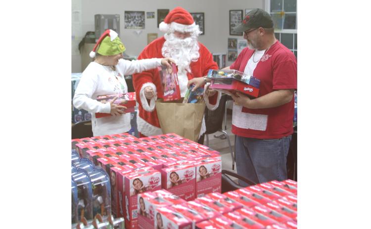 Kimberly Belcher (from left), Santa, and John Cole pack a bag with toys during the Kids Feeding Kids Grinch Christmas event on Sunday evening. (TONY BRITT/Lake City Reporter)