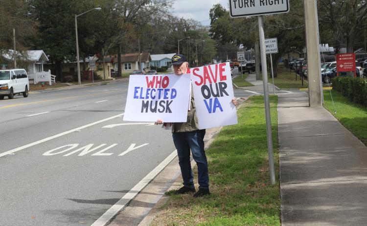 Charles Trowbridge, a U.S. Navy veteran and Fort White resident, holds up signs outside the Lake City VA Medical Center on Monday protesting the Department of Government Efficiency’s planned cutting of 80,000 Veterans Affairs employees. (TONY BRITT/Lake City Reporter)