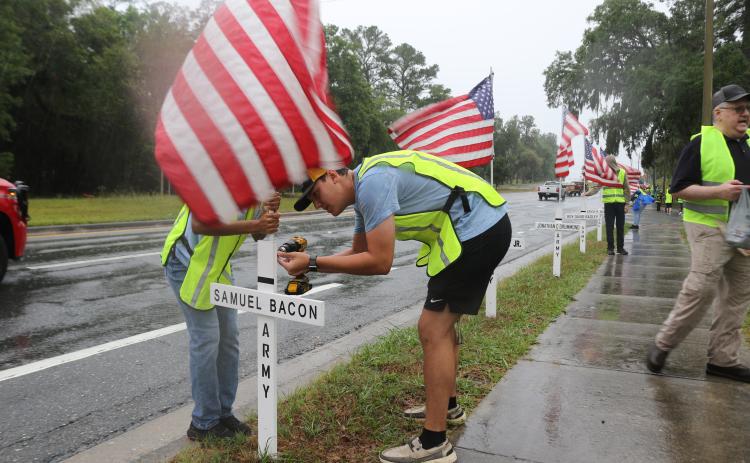 Chase Piesel (right) uses a drill to secure a flag to a cross on Saturday morning as volunteers erected flags in honor of veterans and first responders. (TONY BRITT/Lake City Reporter)
