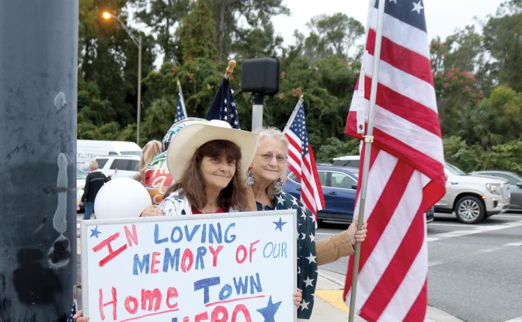 Bonnie Smith (from left) and Doreen Waters, the late Terry Hancock’s sisters, hold flags and a sign for motorists in honor of their brother on Wednesday morning. (TONY BRITT/Lake City Reporter)