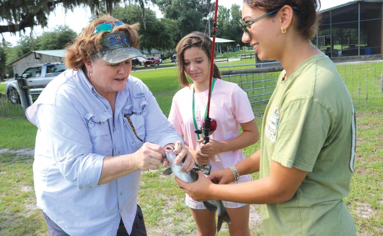 Paulette Tomlinson (from left), a Columbia County Extension Service employee, takes the hook out of a catfish for Madelyn Bullock, 13, with help from Paola Ortiz, a 4-H summer camp volunteer. (TONY BRITT/Lake City Reporter)