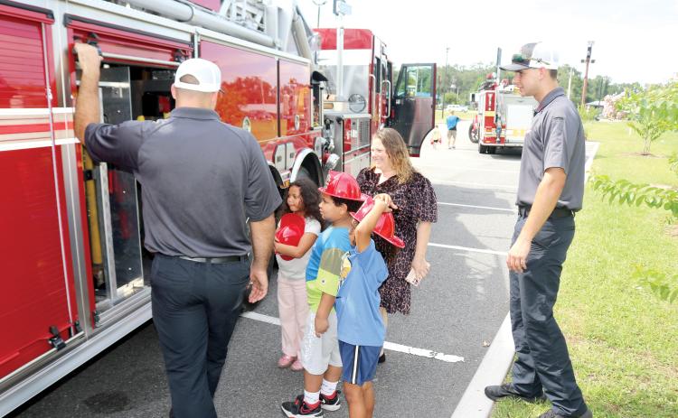 Lake City Fire Department firefighter Chase Hancock (from left) lets Ryker Paris, 7, Jeremiah Williamson, 7, Ariella Paris, 7 and Melissa Angeles look at the equipment firefighters use on emergency calls during the Firefighters and Cones event. (TONY BRITT/Lake City Reporter)