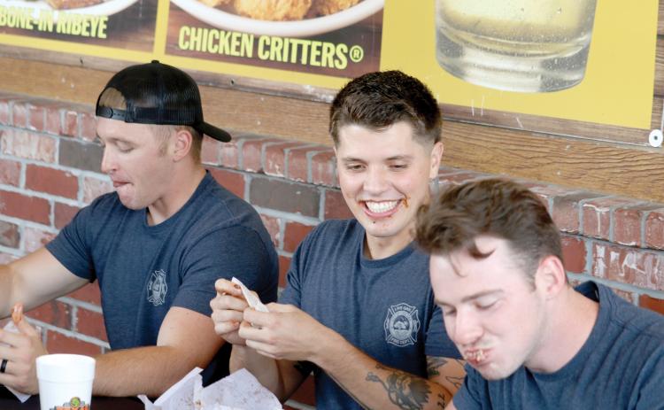 Live Oak firefighter Zach Yarnell (center) grins as Cody Byers (right) has a mouthful of ribs during the contest Tuesday. Kessler Mallory (left) also participated for the Live Oak Fire Department team. (TONY BRITT/Lake City Reporter)