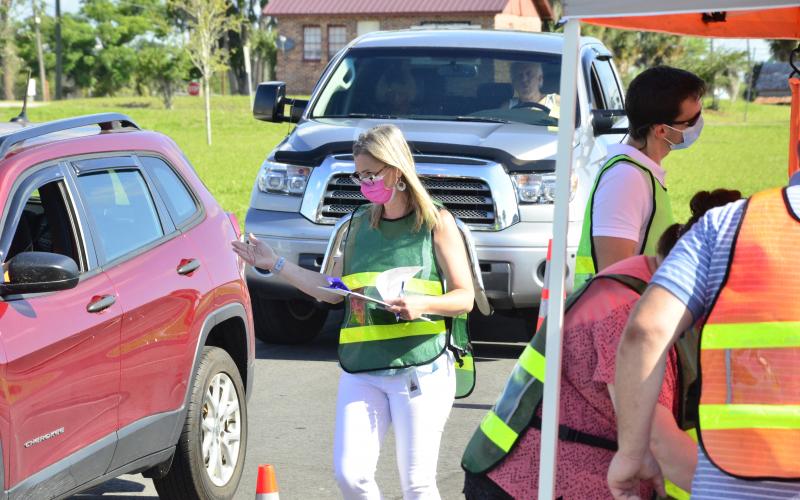 A health official directs traffic during a drive-thru covid-19 testing event Tuesday morning at Wilson Park by Lake DeSoto. CENTER: Workers from the Florida Department of Health suit up to begin testing. (CARL MCKINNEY/Lake City Reporter)