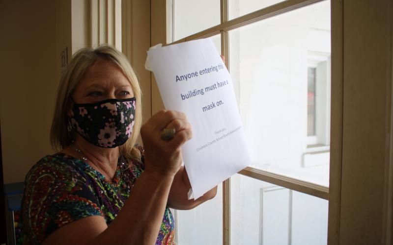 Marsha Wallace, Columbia County School Board office information specialist, places a sign on the central office’s door notifying the general public they must wear masks before entering the building. (TONY BRITT/Lake City Reporter)