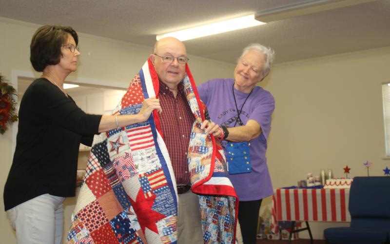 Terry Demott (center), a US Army veteran who served in the Vietnam War, is draped by Dianne Shepherd and Fran Vogt on Wednesday morning at a Quilts of Valor ceremony.  The quilts were made by the Lady of the Lake Quilt Guild members. (TONY BRITT/Lake City Reporter)
